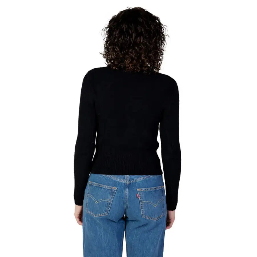 Person with curly dark hair in a black top and blue jeans, modeling Guess Women’s Knitwear
